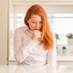 Redhead woman at kitchen feeling unwell and coughing as symptom for cold or bronchitis. Healthcare concept.