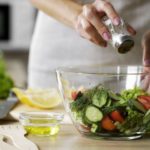 Woman adding salt in vegetable salad glass bowl, health care, excessive salting
