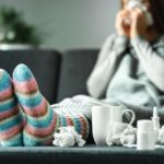 Sick woman with flu, cold, fever and cough sitting on couch at home. Ill person blowing nose and sneezing with tissue and handkerchief. Woolen socks and medicine. Infection in winter.