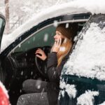 Young woman calling for help or assistance inside snow covered car. Engine start in frost. Breakdown services in the winter