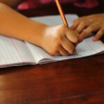 Young hand writing a lesson with a pencil on a book on wooden table – educational concept