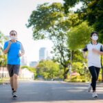 Asian young couple woman and man are jogging and exciseing outdoor in city park and wearing protective mask on face for stay in fit during Covid-19 pandemic in Bangkok, Thailand.