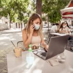 Young woman wearing protective face mask and using hand sanitizer while working remotely on computer laptop in coffee shop outdoors. The New Normal and government health regulations against COVID-19.
